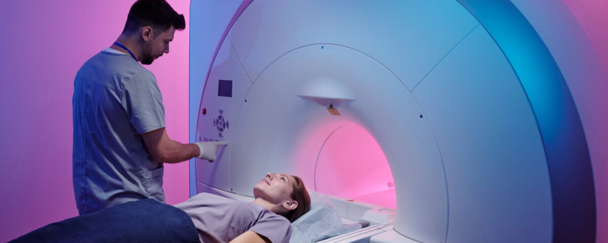 A medical technician operates an MRI machine as a patient lies on the scanning table in a room with pink and blue lighting.