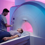 A medical technician operates an MRI machine as a patient lies on the scanning table in a room with pink and blue lighting.