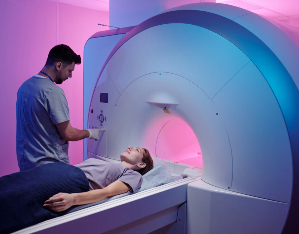 A medical technician operates an MRI machine as a patient lies on the scanning table in a room with pink and blue lighting.